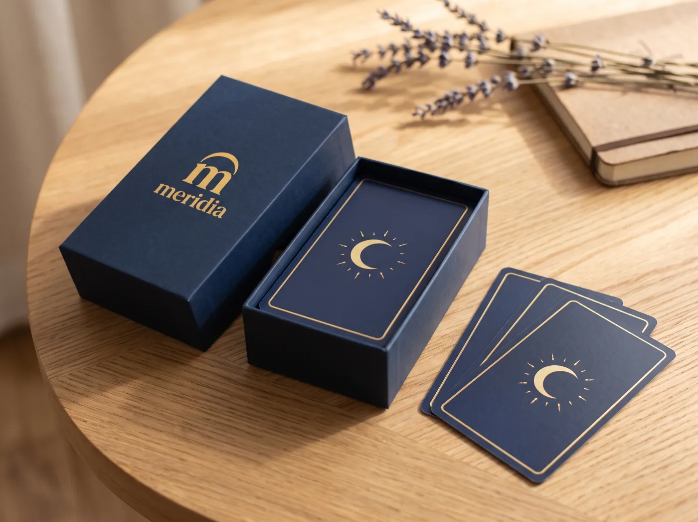 La caja azul de Meridia abierta, con el mazo de cartas adentro, tres cartas al lado y un cuaderno sobre una mesa de madera